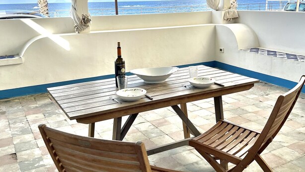An outdoor patio with a table and two wooden chairs, overlooking the ocean.