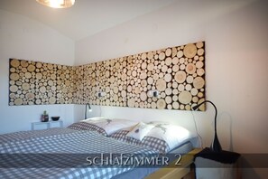 Een slaapkamer met een geruite sprei, een houten kunstwerk van houtblokken aan de muur, een nachtkastje met een lamp en een bijzettafeltje met een plant.