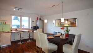 A modern kitchen with wooden flooring, a dining area with a dark wooden table and white chairs, and a large window offering a view of the outdoors.