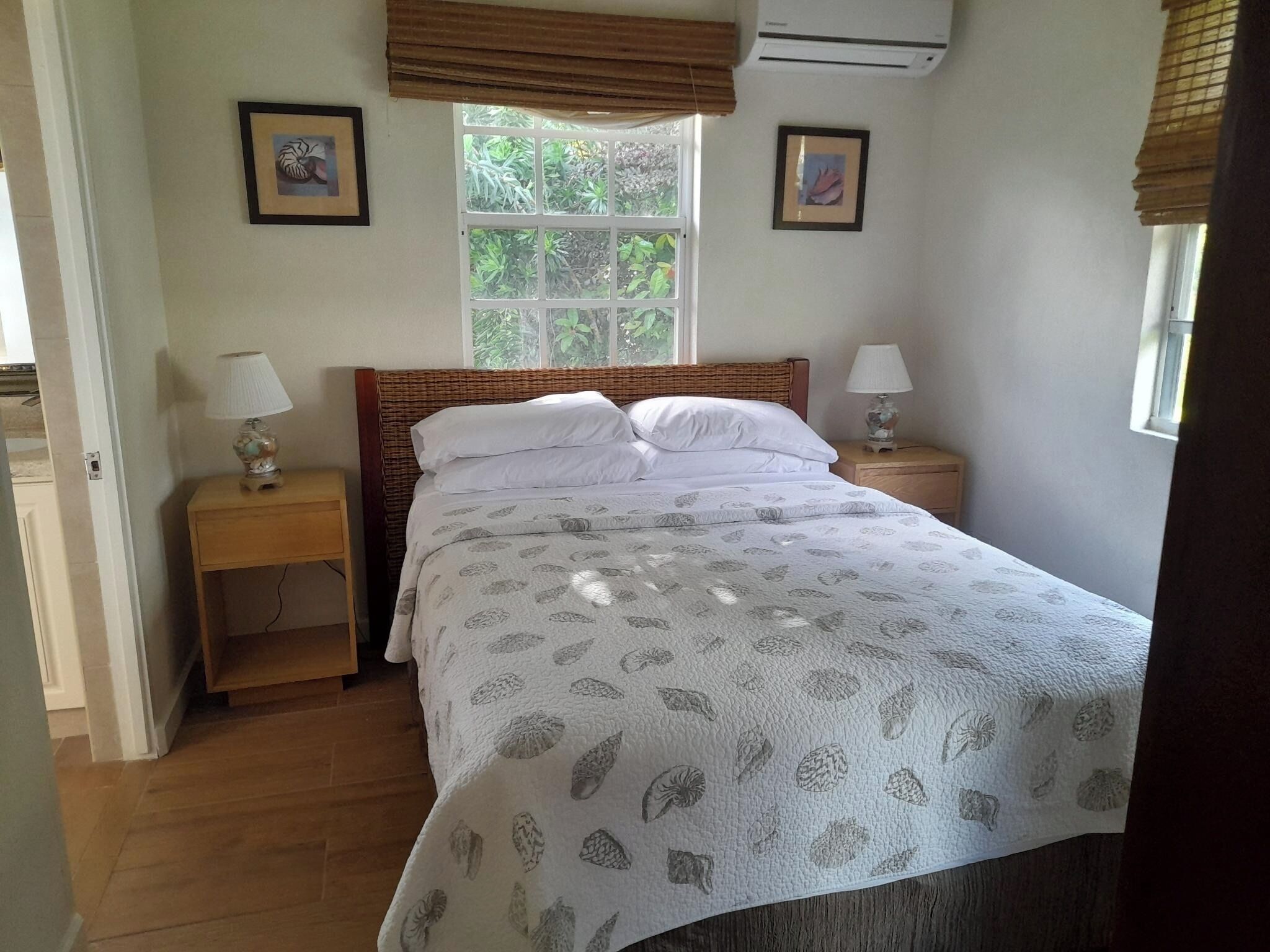 Une chambre à coucher comprenant un lit, deux tables de chevet, une fenêtre donnant sur un paysage verdoyant et deux tableaux accrochés au mur.