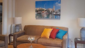 Ein Wohnzimmer mit einem braunen Sofa, bunten Kissen, einem Glastisch und einem Stadtbild an der Wand.