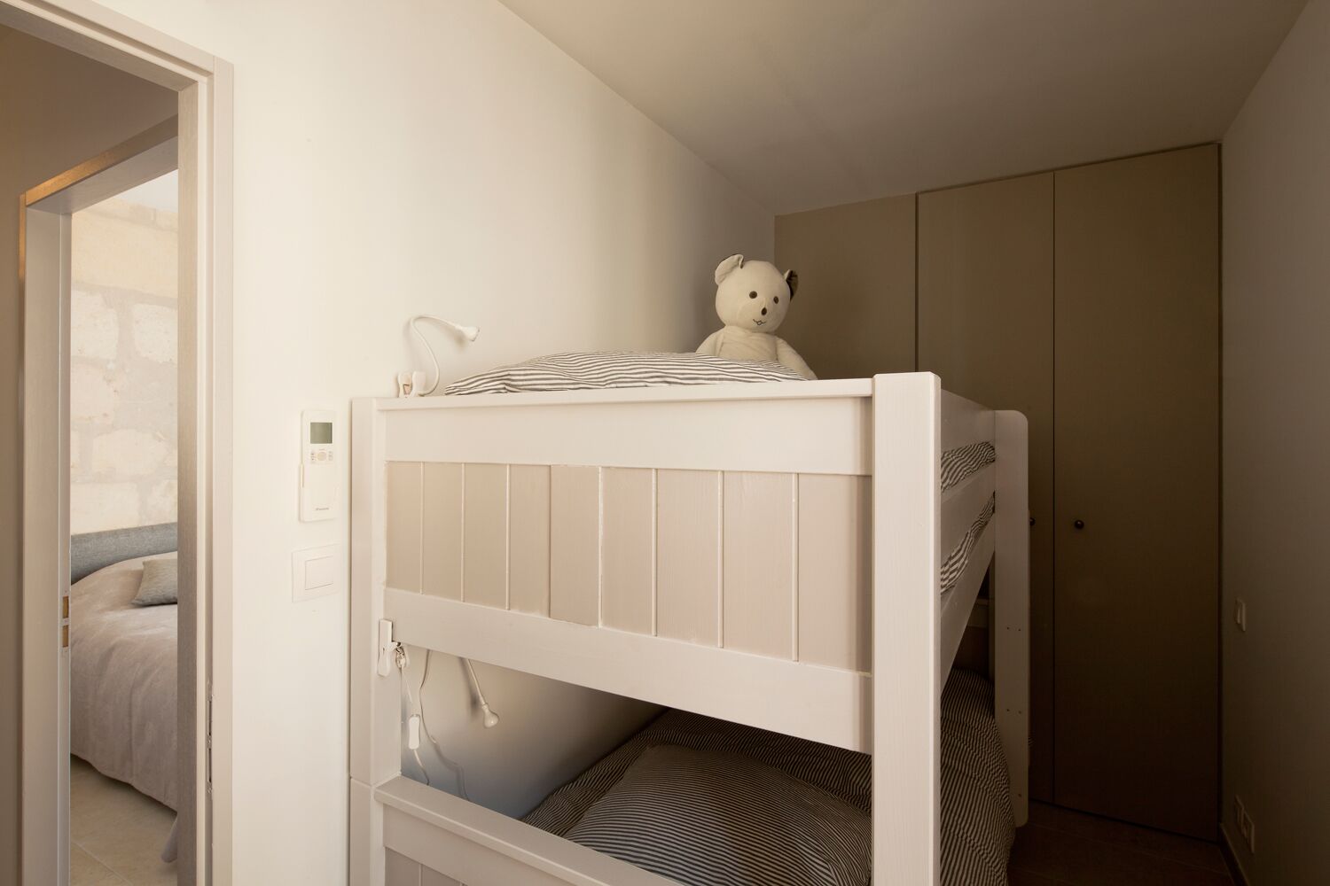 Een stapelbed met een wit hoofdbord, een teddybeer bovenop en een gestreept kussen.