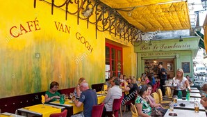 Um café com paredes amarelas e detalhes vermelhos, mesas ao ar livre e um quadro de menu visível.