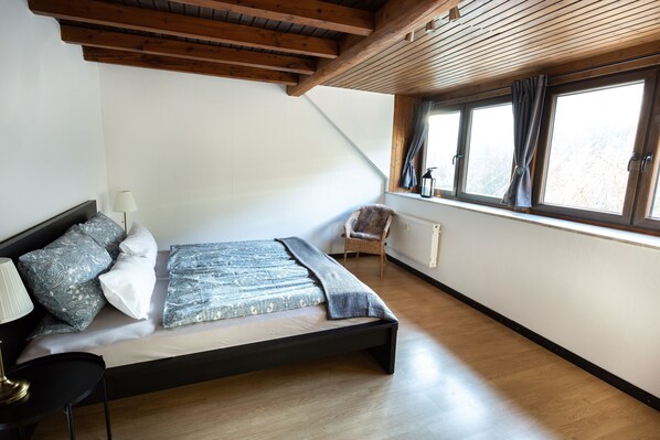 A bedroom with a bed, a chair, a window, and wooden beams on the ceiling. - Country Living in the southern Eifel, vacation apartment in the Sauertal N° 2 (Ralingen)