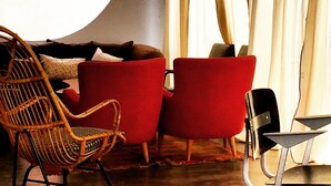 Un salon moderne avec un grand luminaire rond, un fauteuil en osier, deux fauteuils rouges et une petite table d’appoint.
