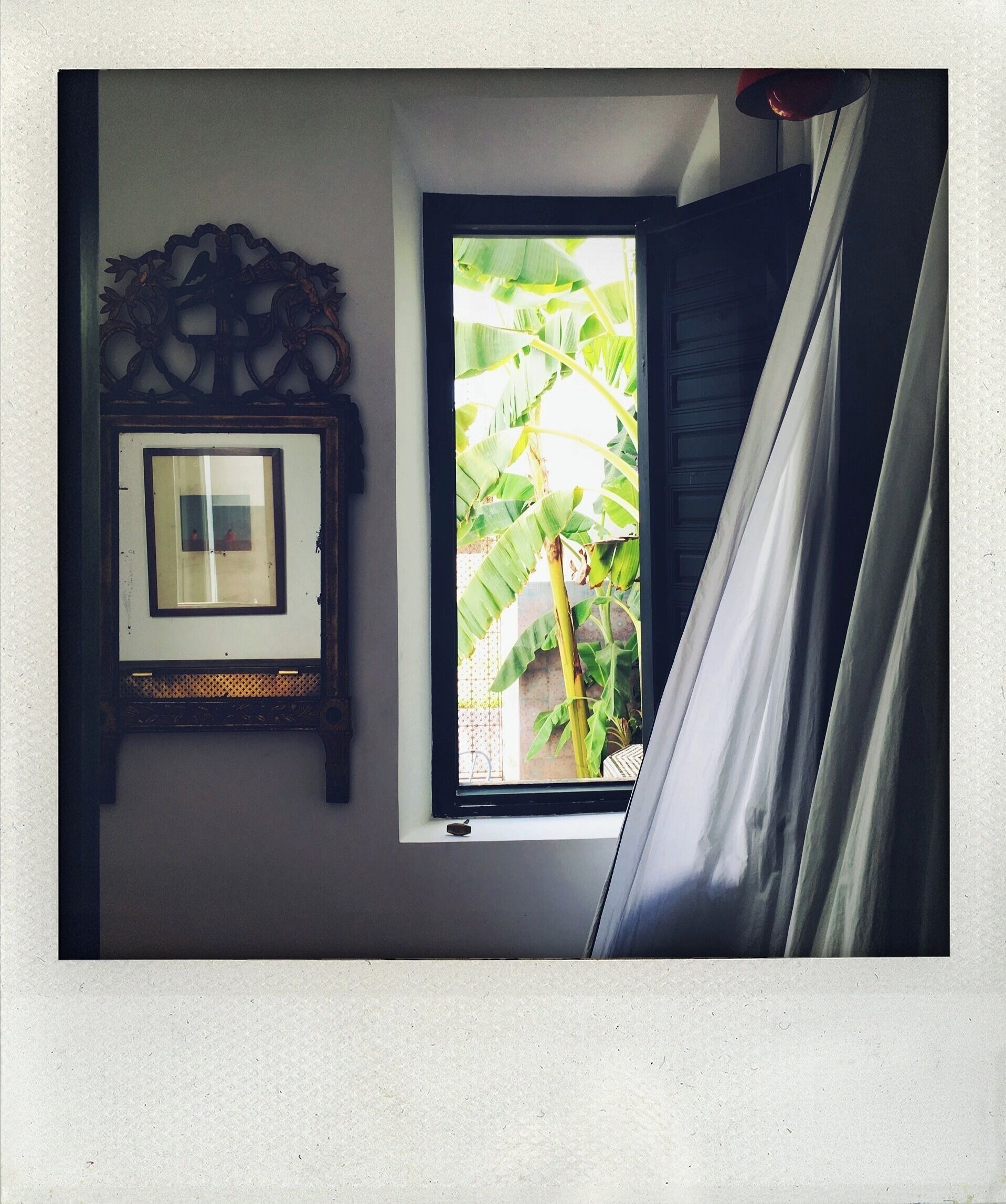 Quarto com um quadro na parede, janela com vista para bananeira e cortina entreaberta.