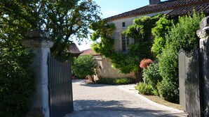 A courtyard with a gravel path leading to a gated entrance, surrounded by lush greenery and a building with a tiled roof.