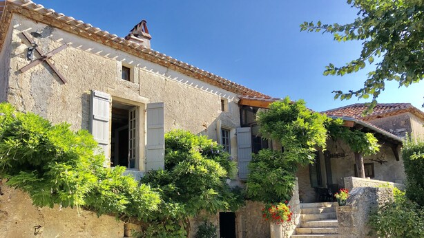 A rustic stone building with a tiled roof, a wooden staircase, and lush greenery.