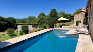 A rectangular swimming pool with a stone deck, surrounded by a grassy area and trees.
