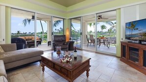A spacious living room with a large glass door leading to an outdoor area, a ceiling fan, and a television on a wooden cabinet. - Partial Ocean Views, Top Floor. Beach Gear Included.  Vista Waikoloa A304 (Waikoloa Beach Resort)