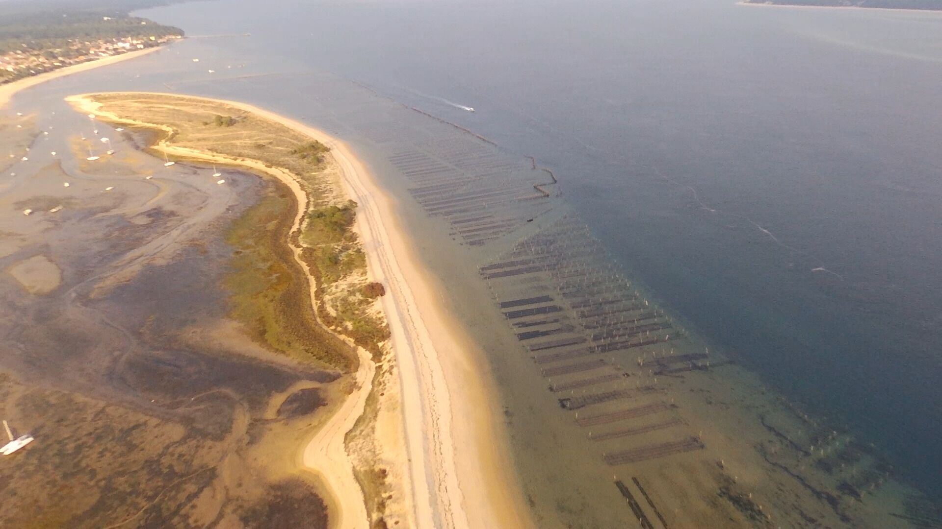 An aerial view of a coastal area with a sandy beach, a marina with boats, and a distant shoreline with buildings.