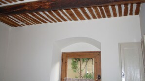Ein Zimmer mit weißen Wänden, einer Holzbalkendecke, einer Tür und einem Fenster mit Blick auf Grünflächen.