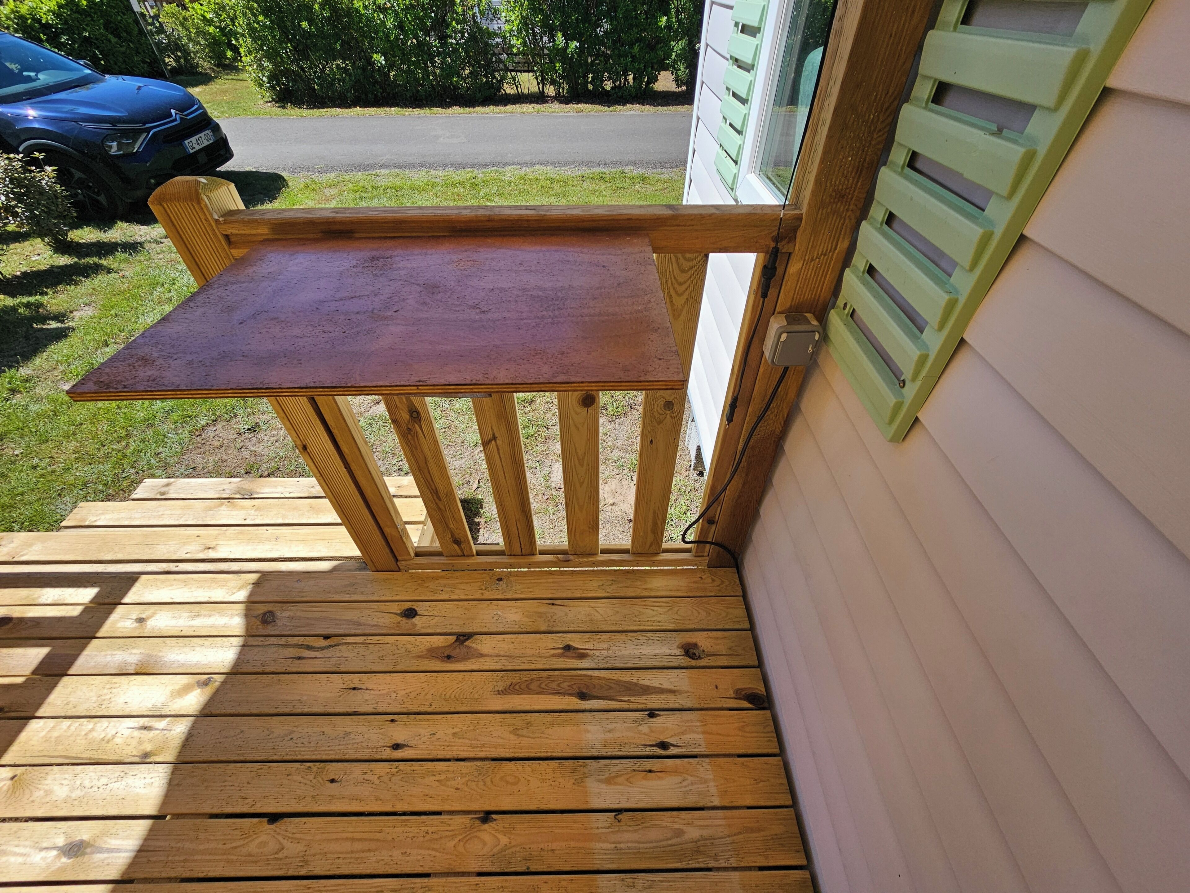 Una terraza de madera con una pequeña mesa y una silla, que da a una casa con contraventanas verdes.
