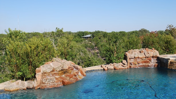 A swimming pool with natural rock formations and a view of a hill with a building. - Love of the View  - Glen Rose Luxury Cottage (Glen Rose)