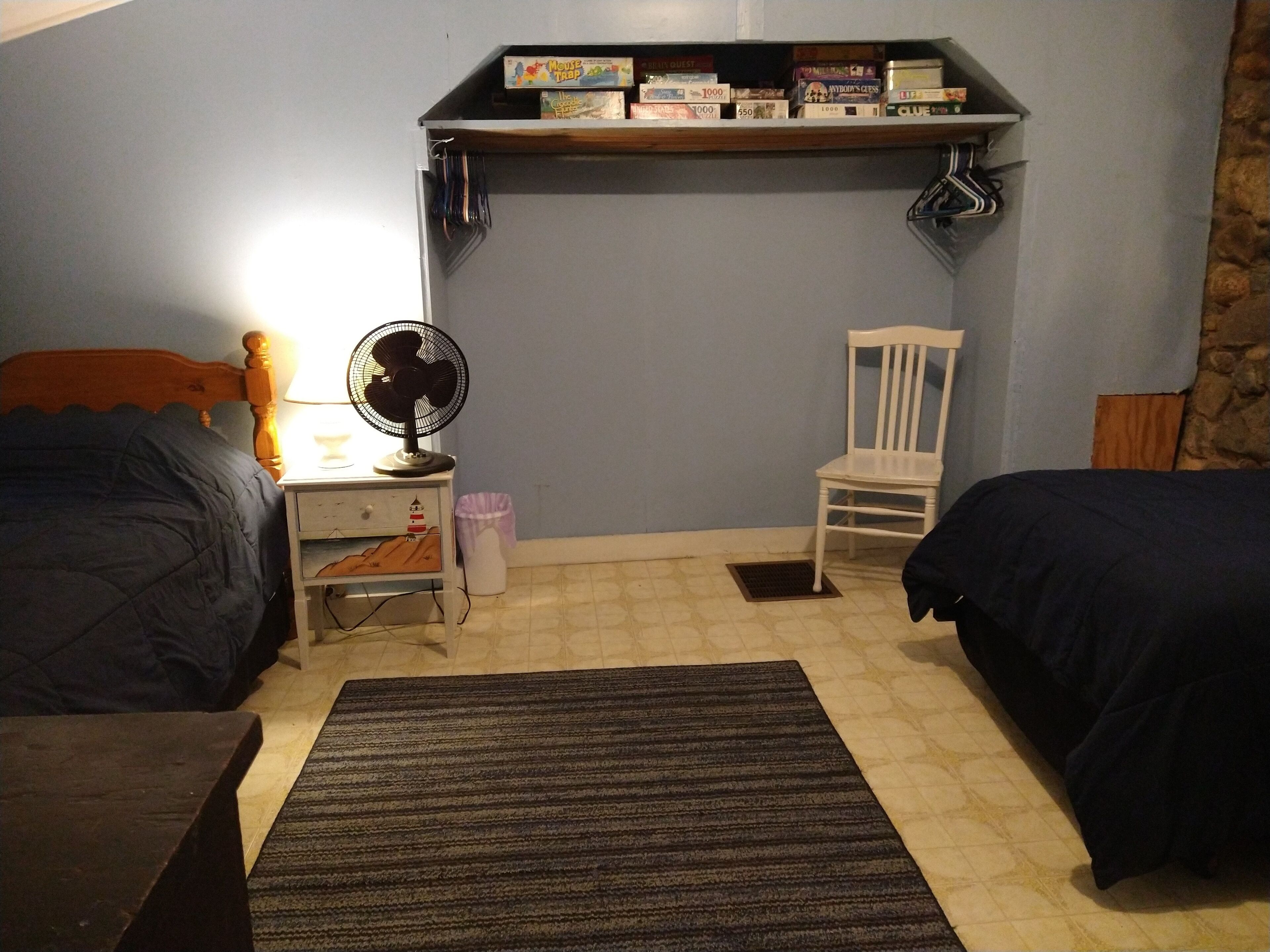 A bedroom with two beds, a nightstand, a chair, a shelf with board games, and a fan.