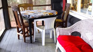 A wooden table with a floral tablecloth, surrounded by wooden chairs, a wicker sofa with a red cushion, and a window with a view of trees. - Rustic two-story log cottage, River-view, Pet-friendly, Downeast Maine (Edmunds Township)