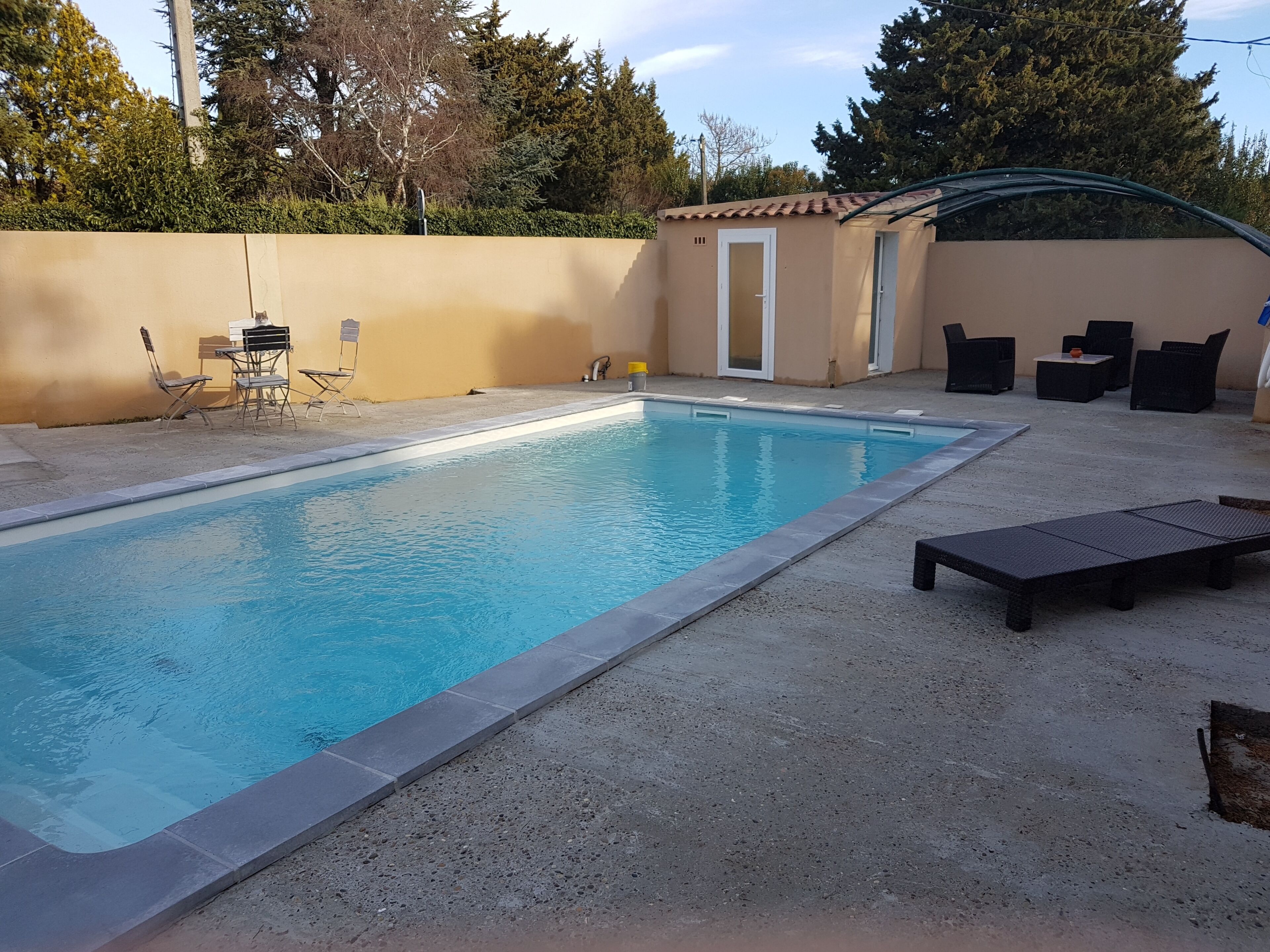 Une piscine rectangulaire bordée de carreaux, entourée d’une terrasse en béton. On y trouve des chaises longues et une petite table près de la piscine.