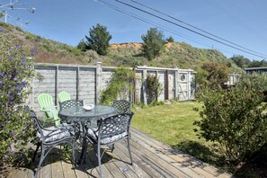 A wooden deck with a glass table and chairs, surrounded by greenery and a hillside in the background. - Mermaid's House (Admiral's Quarters) - Tomales Bayfront, Point Reyes, Marshall (Marshall)