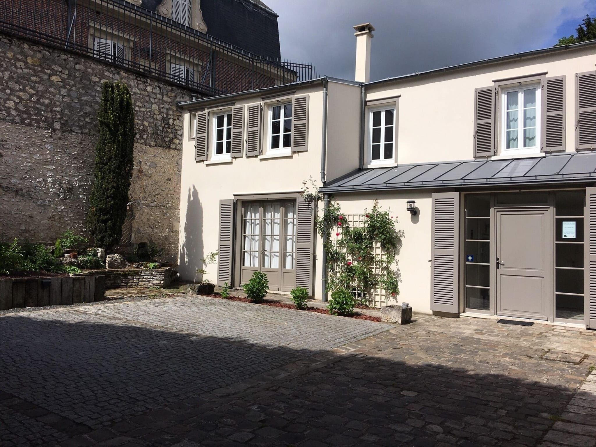 A two-story building with a stone facade, shutters, and a paved courtyard.