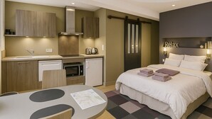 Quarto de hotel compacto com cozinha compacta, uma cama com almofadas, uma pequena mesa com cardápio e um letreiro com a palavra 'Relax' na parede.