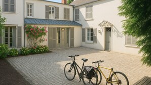 Um pátio com duas bicicletas, um edifício branco com persianas e um pequeno jardim com flores cor-de-rosa.