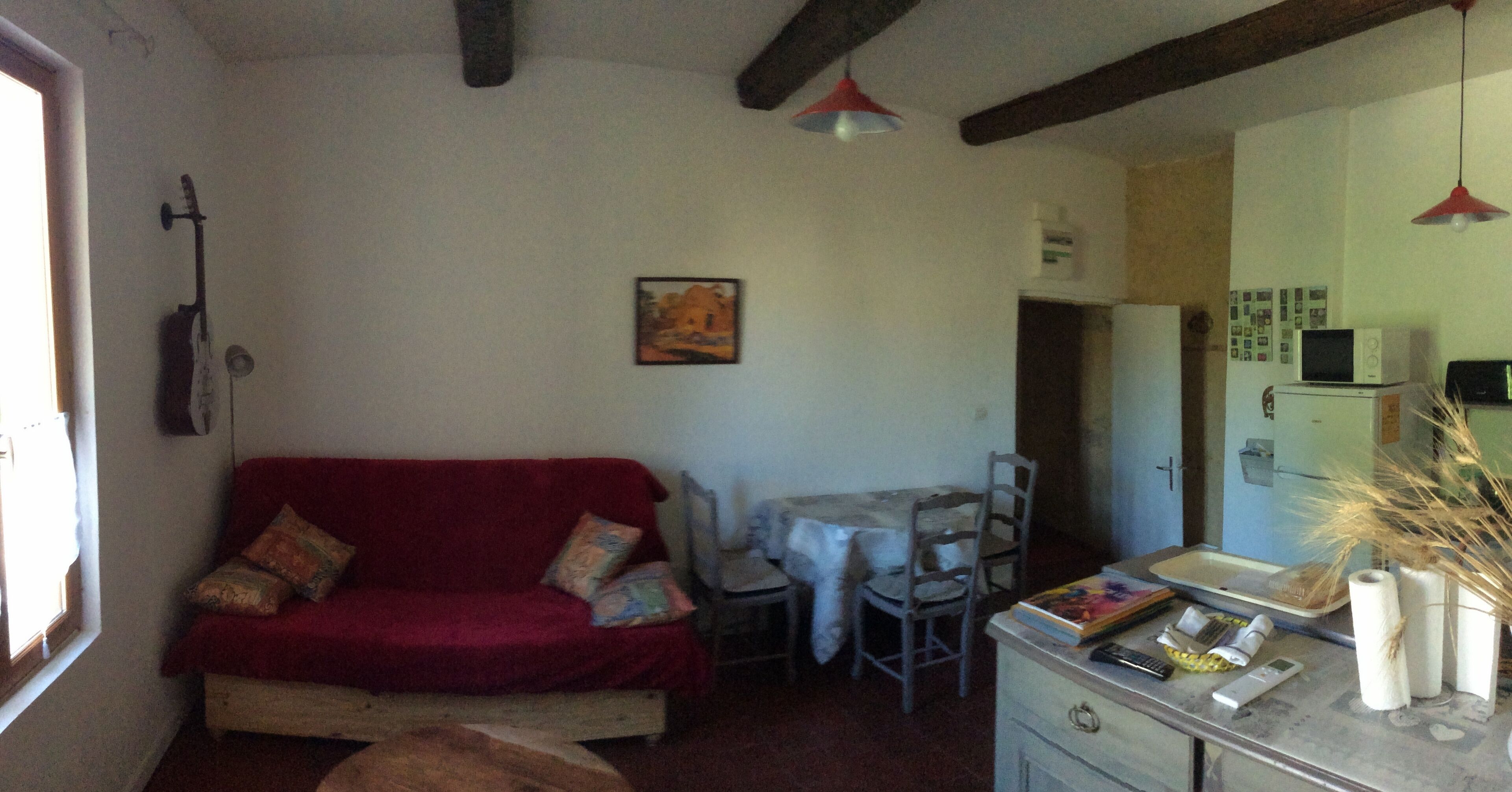 A cozy living room with a red sofa, a dining table with chairs, a kitchen area with a microwave, and a painting on the wall.