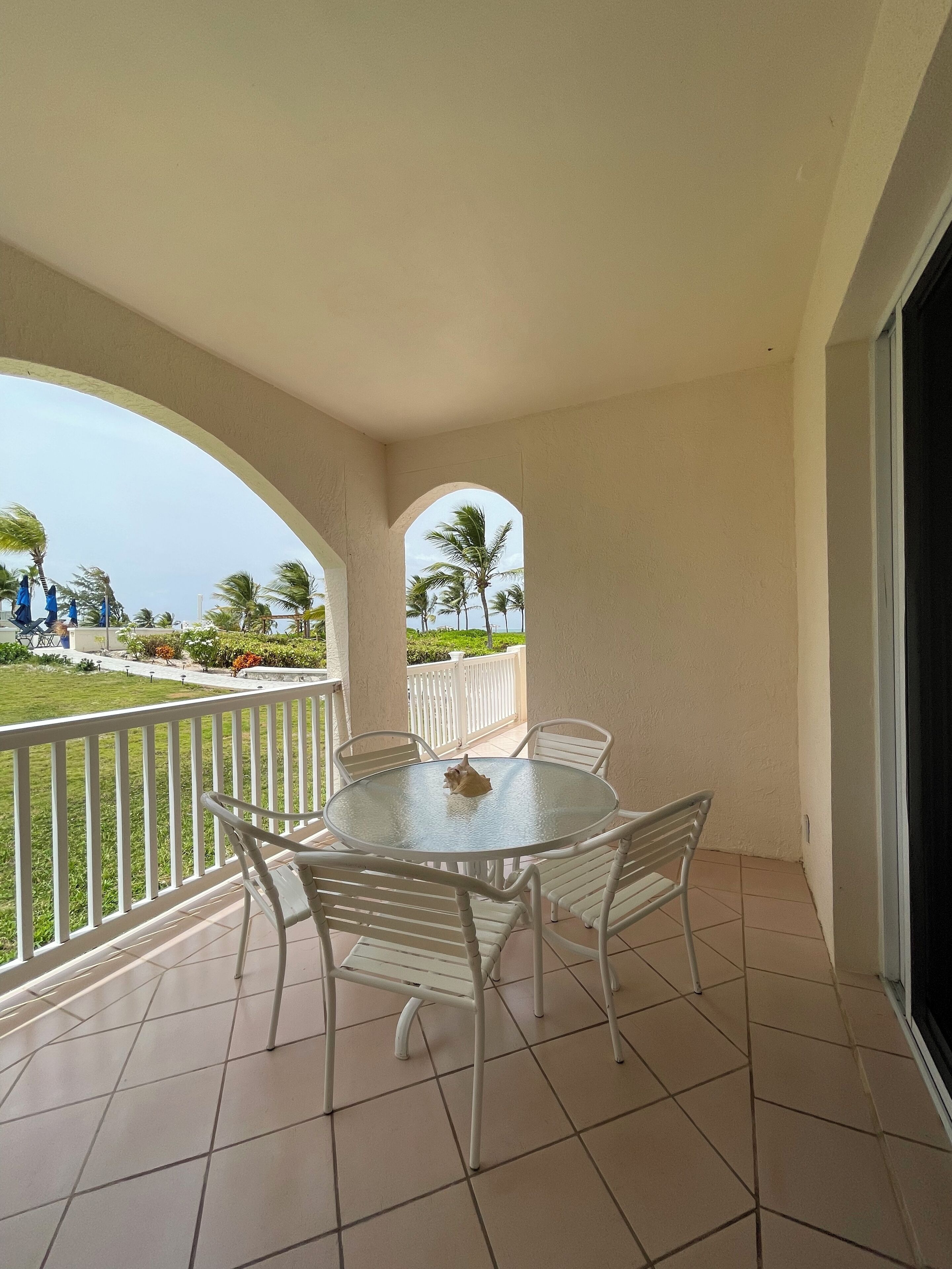 A balcony with a round table and chairs, a view of a lawn and palm trees, and a white railing.