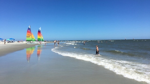 Una playa con cielos despejados, una orilla arenosa y algunas personas y veleros.