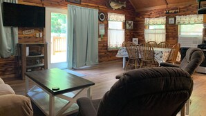 Interior rústico de una cabaña con una mesa de comedor, sillas, un televisor y paredes de madera.