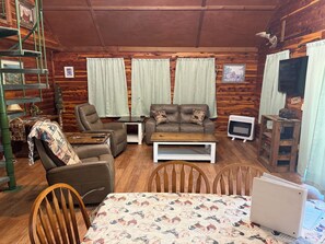 A cozy cabin interior with a sofa, armchairs, a dining table, and a television.