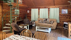 Interior acogedor de una cabaña con una escalera de caracol, paredes de madera, un sofá, una mesa de comedor y un televisor pequeño.