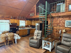 A cabin interior with a dining area, two leather chairs, a coffee table, a kitchen with wooden cabinets, a refrigerator, a stove, and a spiral staircase.