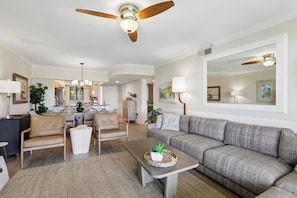 A spacious living room with a ceiling fan, a grey sofa, a coffee table with a plant, and a dining area with chairs and a chandelier.