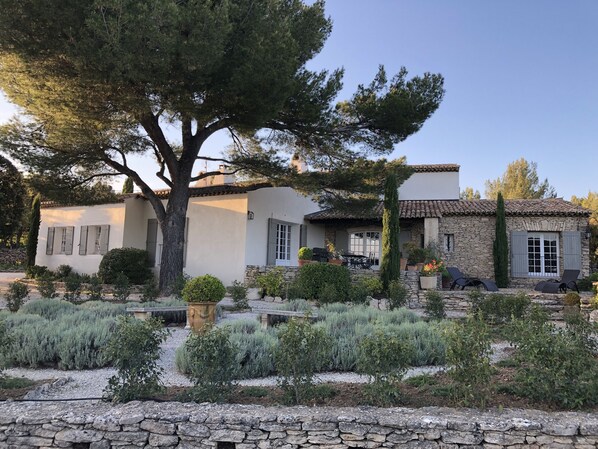 A two-story building with a stone facade, a patio with outdoor furniture, and a garden with various plants. - Gorgeous in Gordes, Elegantly Furnished with Pool, Tennis and Views  (Gordes)