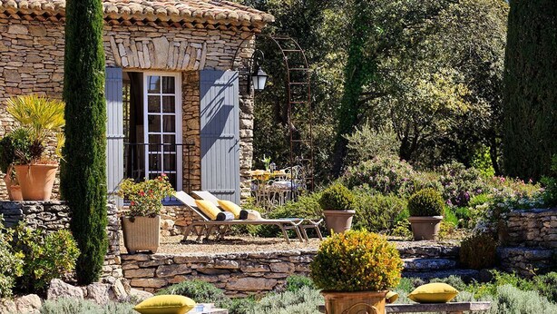 A stone house with a tiled roof, a wooden door, and a small patio with lounge chairs and potted plants. - Gorgeous in Gordes, Elegantly Furnished with Pool, Tennis and Views  (Gordes)