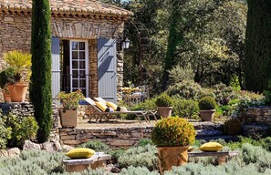 A stone house with a tiled roof, a wooden door, and a small patio with lounge chairs and potted plants. - Gorgeous in Gordes, Elegantly Furnished with Pool, Tennis and Views  (Gordes)