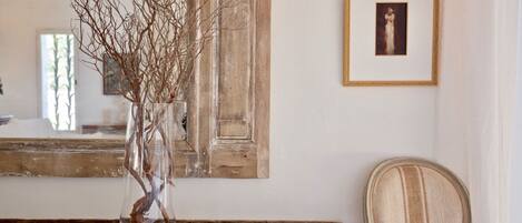 A rustic wooden table with a clear glass vase containing branches, a framed picture on the wall, and a wicker chair.