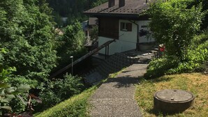 Een landhuis met een stenen pad dat naar de ingang leidt, omringd door weelderig groen en bergen op de achtergrond.