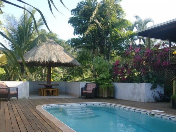 A swimming pool with a wooden deck, a thatched roof structure, and lush tropical vegetation.
