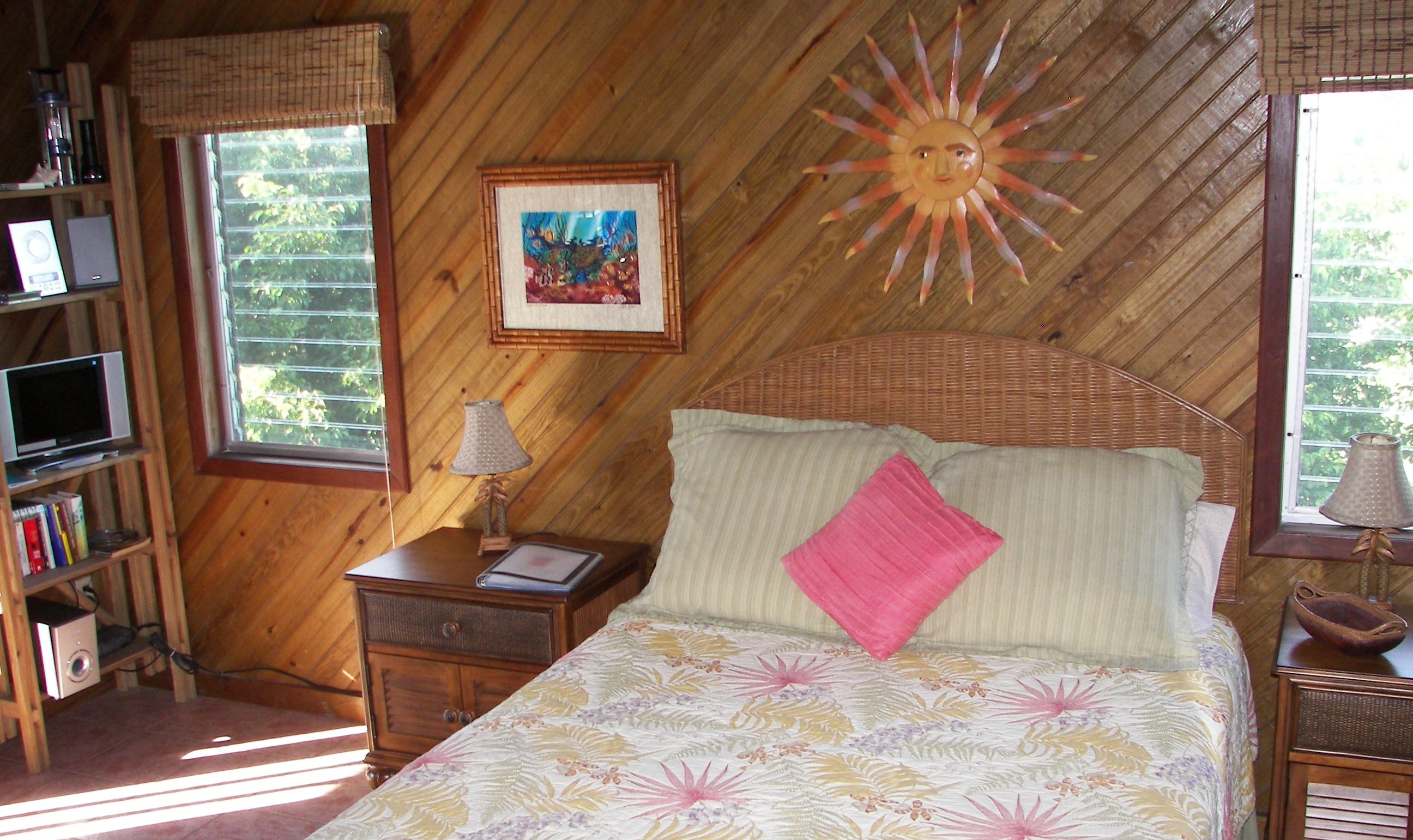 Een slaapkamer met een bed, nachtkastje, boekenkast en raam met zon decoratie.