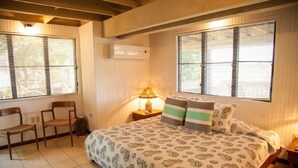 A bedroom with a bed, two chairs, a nightstand with a lamp, and a window with blinds. - Punta Aloe 20 Ocean Front Villa Apartment (Culebra)