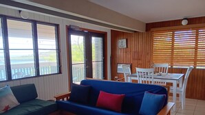 A room with a blue sofa, a wooden dining table with white chairs, and large windows with wooden blinds. - Punta Aloe 20 Ocean Front Villa Apartment (Culebra)