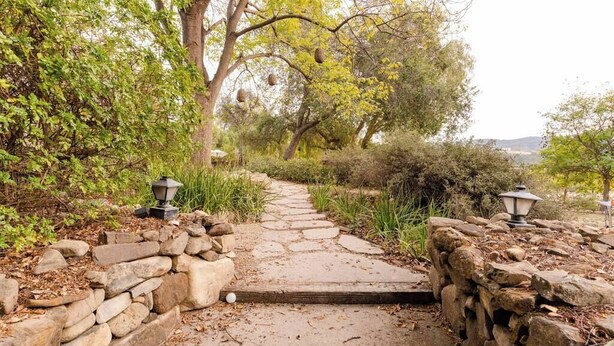 A stone pathway leading through a garden with trees and stone walls, flanked by lantern-style lights.
