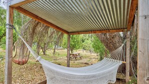 A hammock under a wooden pergola with a corrugated metal roof, surrounded by trees and a picnic table.