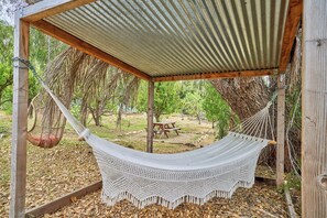 Uma rede sob uma pérgola de madeira com telhado de chapa metálica, cercada por árvores e uma mesa de piquenique.