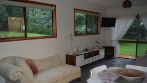 Uma sala de estar com sofá, mesa de jantar com cadeiras, lareira e janelas amplas com vista para o exterior.
