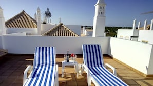 Two striped lounge chairs on a tiled terrace with a table and wine glasses, overlooking a cityscape with white buildings and a clear sky.
