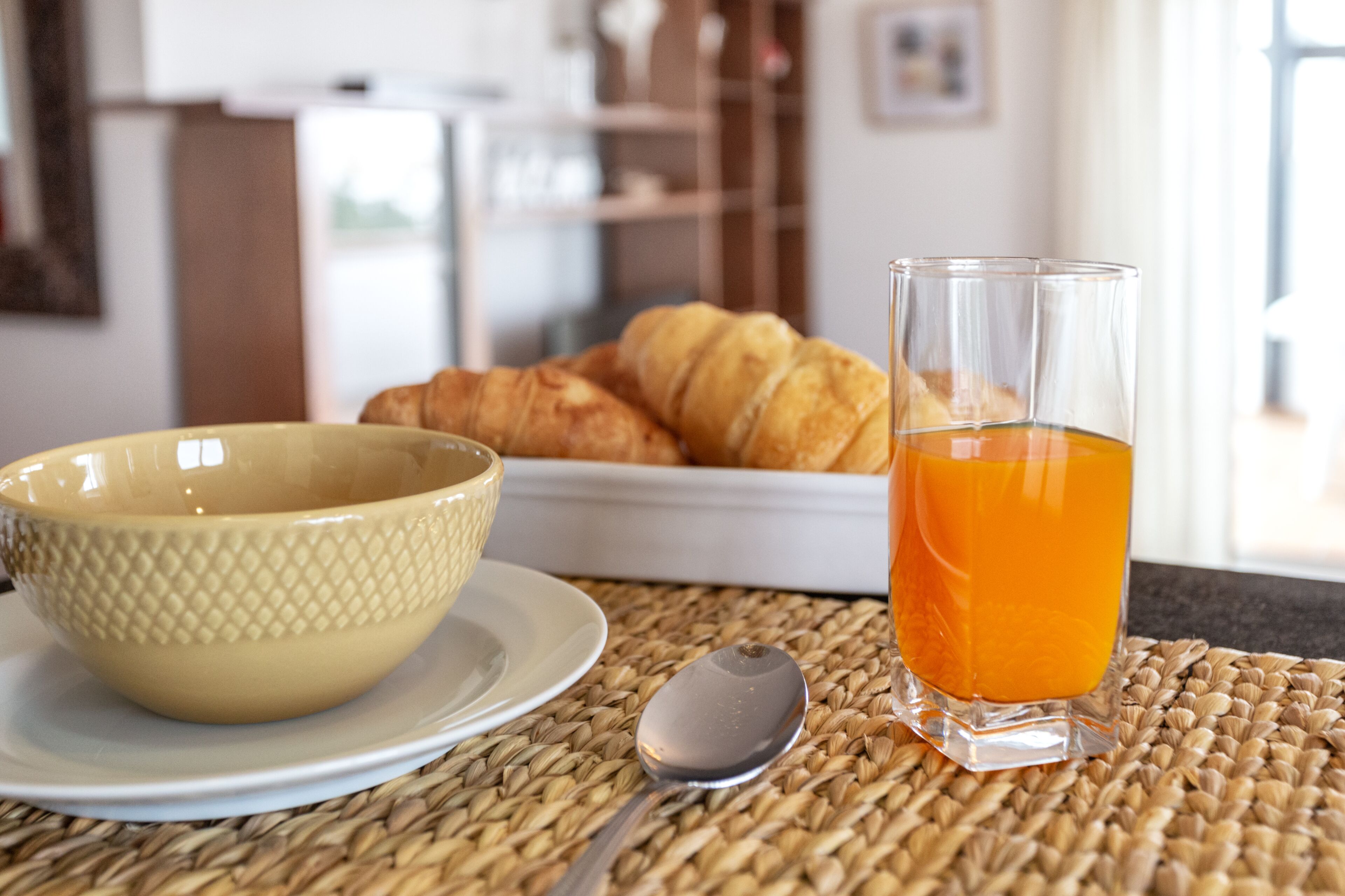 A breakfast setting with a bowl, spoon, glass of orange juice, and croissants on a woven placemat.