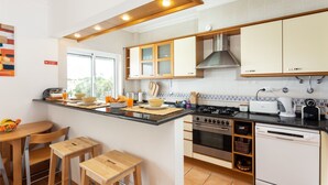 A modern kitchen with wooden cabinets, a gas stove, an oven, and a dishwasher. There is a breakfast bar with chairs and a window providing natural light.