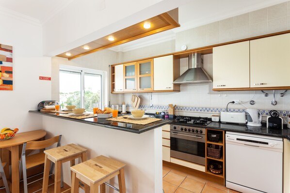 A modern kitchen with wooden cabinets, a gas stove, an oven, and a dishwasher. There is a breakfast bar with chairs and a window providing natural light.